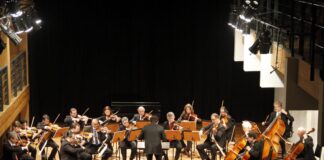 foto mostra os músicos da orquestra de câmara no palco, sendo regidos pelo maestro